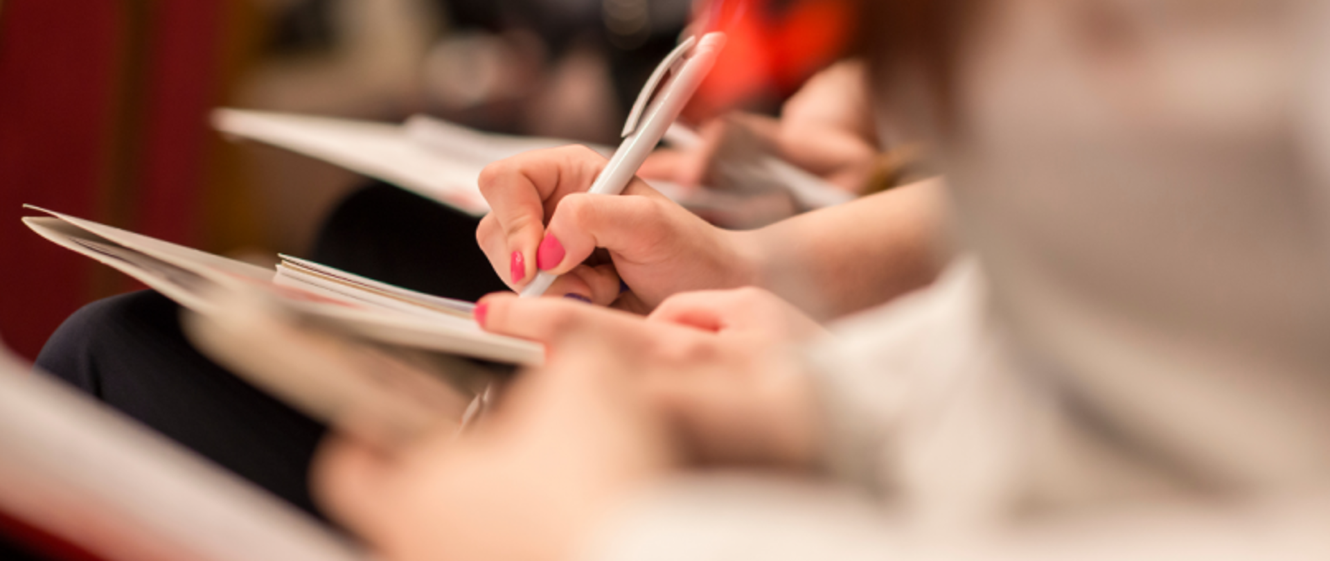 Dłonie osób piszących w zeszytach lub na kartkach, zbliżenie na czynność notowania.