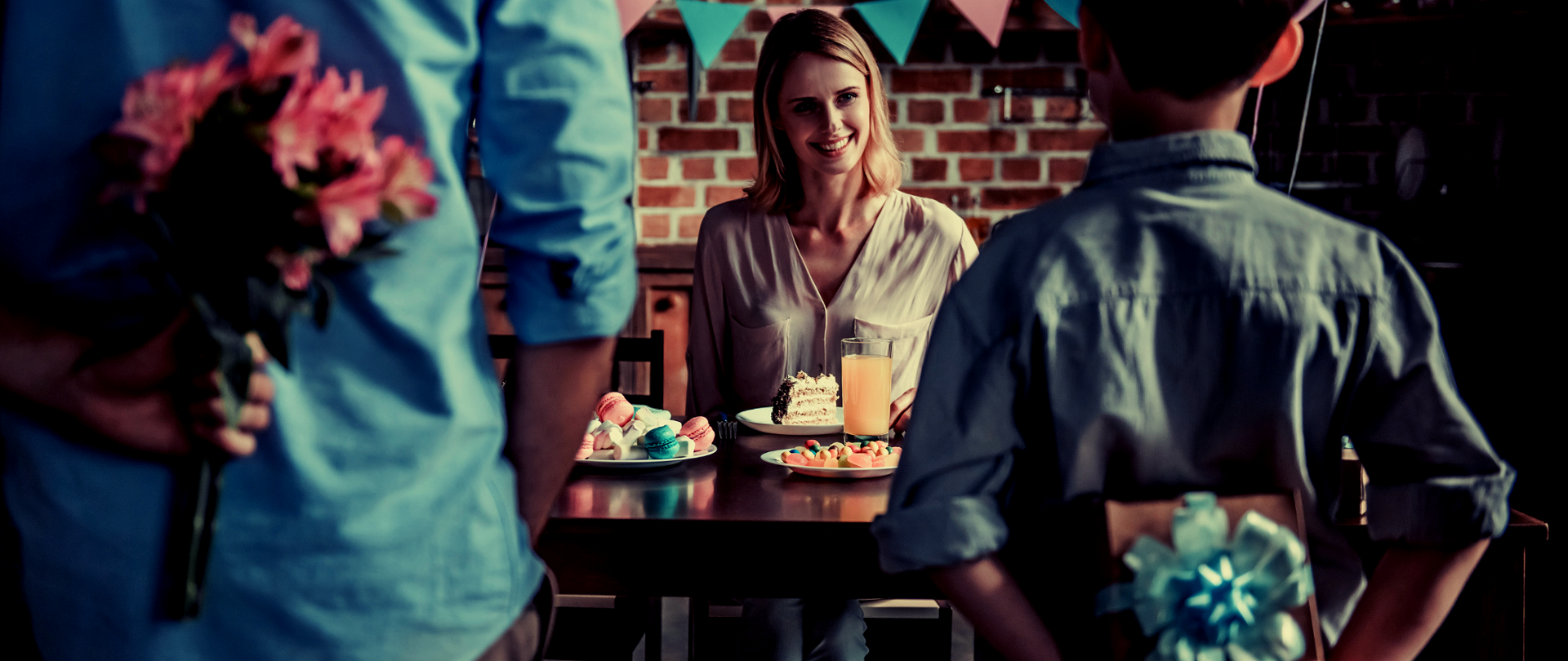 Kobieta siedzi przy stole. Przed nią stoi dwóch chłopców z bukietami kwiatów. 