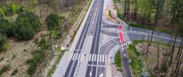 Zdjęcie przedstawia nowoczesną infrastrukturę drogową. Widoczne jest skrzyżowanie w kształcie litery T, na którym zlokalizowano chodnik oraz ścieżkę rowerową. Skrzyżowanie jest skanalizowane i oświetlone. Całość przedstawia nowoczesne rozwiązania komunikacyjne, w którym zrównoważono interesy zmotoryzowanych, pieszych i cyklistów. Skrzyżowanie położone jest w obszarze leśnym. Zdjęcie przedstawia harmonijne połączenie infrastruktury i przyrody.