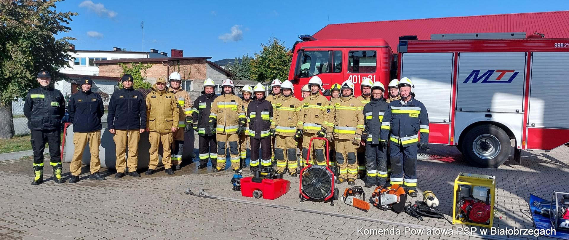 Dziewiętnastu strażaków w ubraniach specjalnych. W tle czerwony samochód pożarniczy, na pierwszym planie sprzęt strażacki (pompy, piły, wentylatory, agregaty.