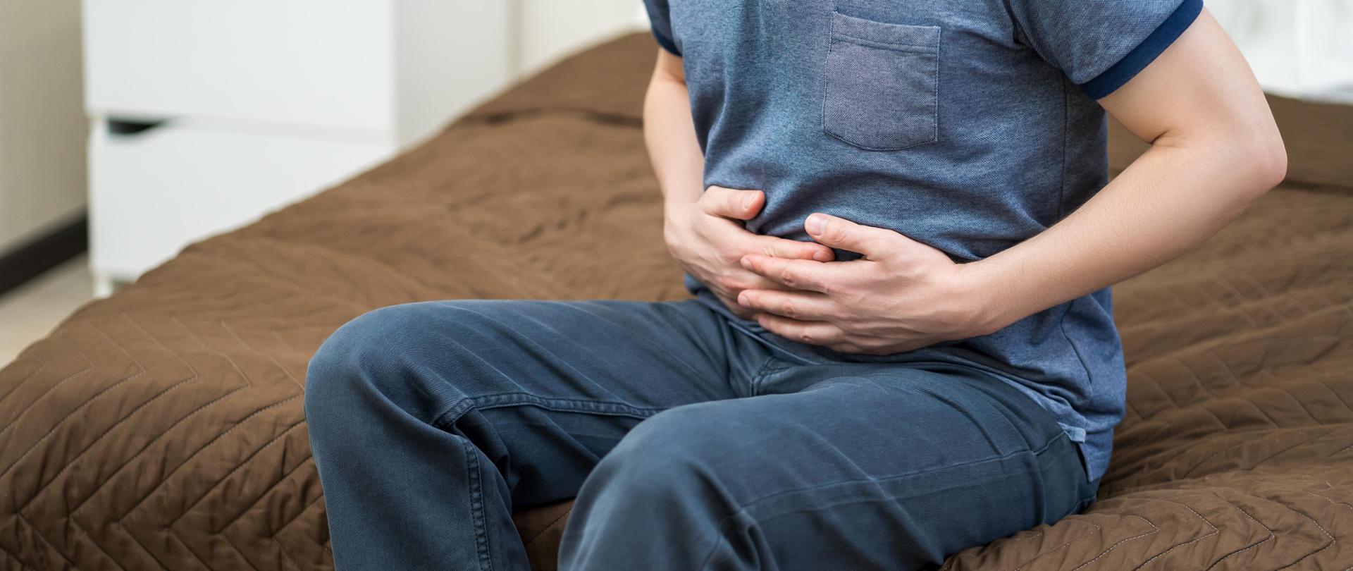 Mężczyzna siedzący na krawędzi łóżka, trzymający ręce na brzuchu.