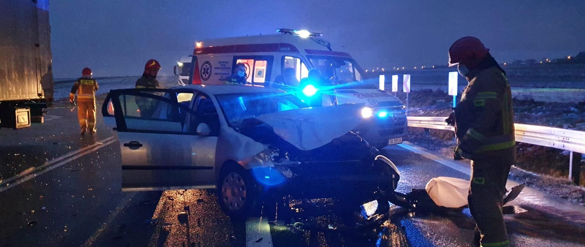 Zdjęcie przedstawia uszkodzony samochód osobowy na drodze wojewódzkiej. W tle widnieje karetka pogotowia z włączoną sygnalizacją alarmową świetlną. Przy samochodzie widać strażaka w umundurowaniu bojowym. Po lewej stronie jeszcze dwóch strażaków. Zdjęcie zrobione w porze nocnej.