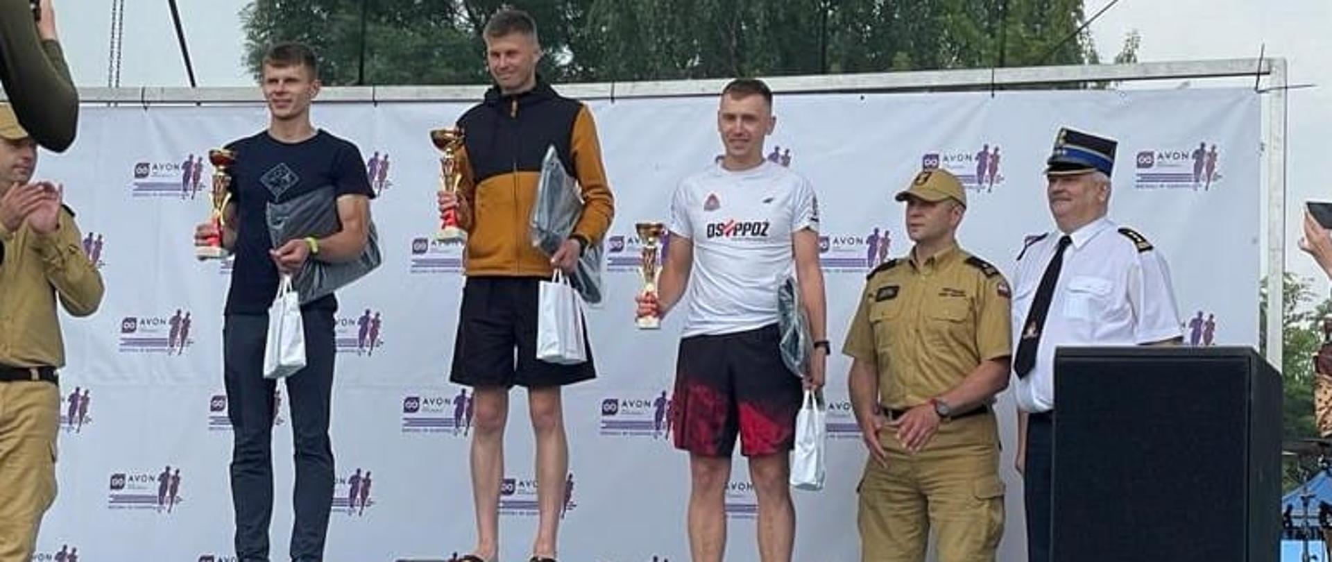 Na zdjęciu widoczne jest 3 osoby stojące na podium obok nich stoją 3 funkcjonariusze PSP.