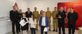 Zdjęcie przedstawia trojkę dzieci – zwycięzców Eliminacji Powiatowych Ogólnopolskiego Turnieju Wiedzy Pożarniczej "Młodzież zapobiega pożarom" – grupa klasy 1-3. Dzieci – dwie dziewczynki i chłopiec – ubrani w stroje galowe stoją z dyplomami i nagrodami dla zwycięzców. Za nimi stoją organizatorzy eliminacji Strażacy PSP i OSP oraz Pan Starosta. W tle elementy dekoracyjne z motywami narodowymi i Państwowej Straży Pożarnej.
