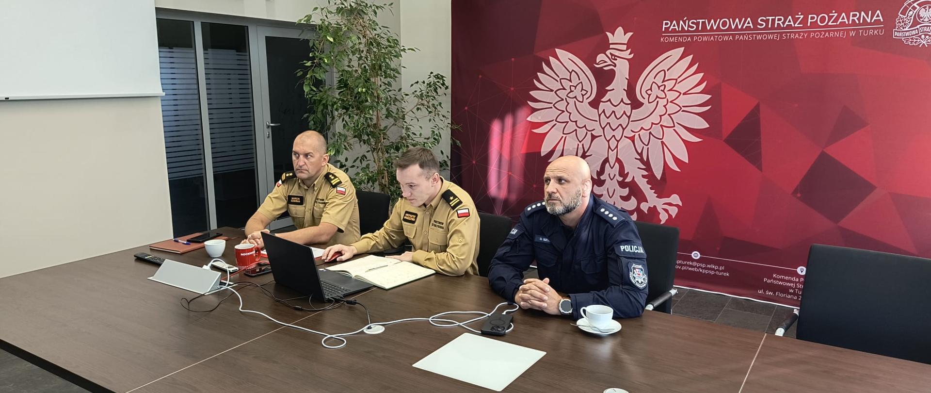 Zbliżenie na trzech uczestników spotkania siedzących z jednej strony stołu konferencyjnego – dwóch strażaków w piaskowych mundurach (jeden z nich obsługuje laptopa) oraz policjanta w ciemnoniebieskim mundurze. W tle widoczna jest ta sama bordowa ścianka z białym orłem Państwowej Straży Pożarnej.