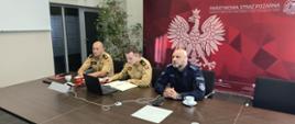 Zbliżenie na trzech uczestników spotkania siedzących z jednej strony stołu konferencyjnego – dwóch strażaków w piaskowych mundurach (jeden z nich obsługuje laptopa) oraz policjanta w ciemnoniebieskim mundurze. W tle widoczna jest ta sama bordowa ścianka z białym orłem Państwowej Straży Pożarnej.