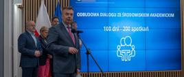 Minister Wieczorek i wiceministrowie stoją z boku sali, pod wielkim wbudowanym w ścianę ekranem z napisem Odbudowa dialogu ze środowiskiem akademickim, minister mówi do mikrofonu na stojaku.