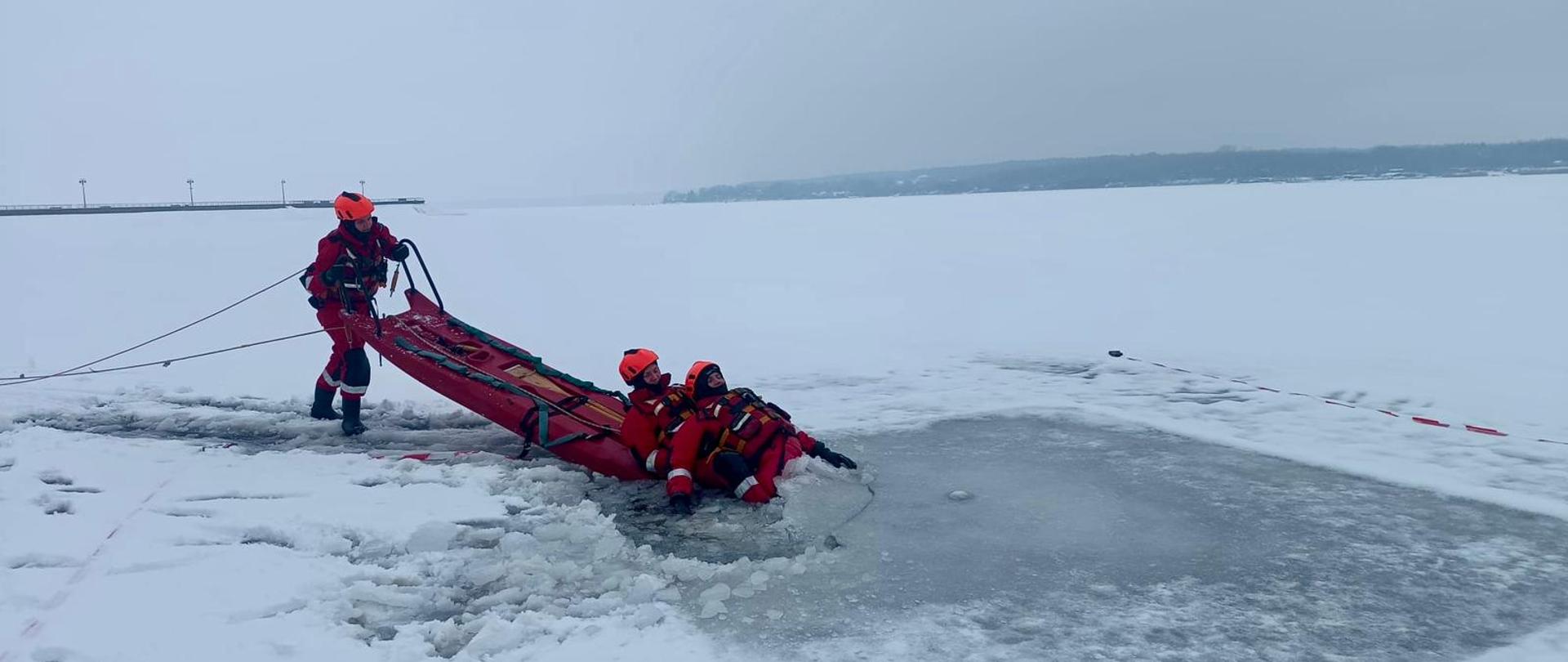 Ratownicy w czerwonych kombinezonach i kaskach prowadzą akcję ratunkową na zamarzniętym jeziorze, wyciągając osoby z przerębla przy użyciu sań lodowych i lin asekuracyjnych.
