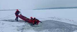 Ratownicy w czerwonych kombinezonach i kaskach prowadzą akcję ratunkową na zamarzniętym jeziorze, wyciągając osoby z przerębla przy użyciu sań lodowych i lin asekuracyjnych.