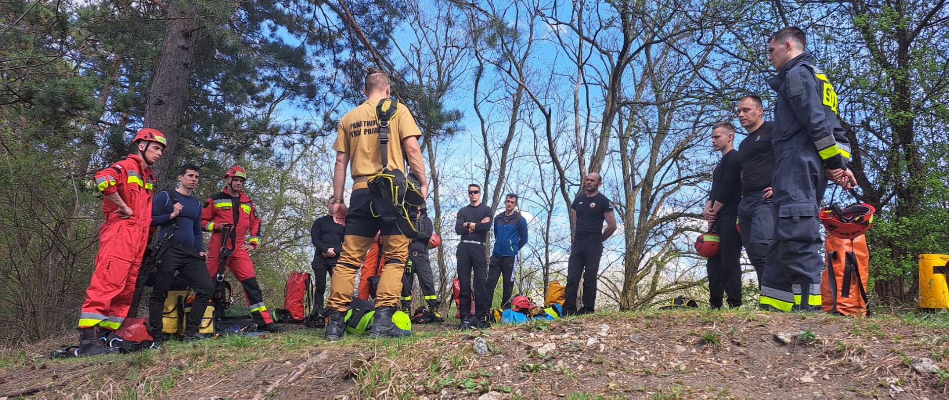 5. Zdjęcie przedstawia grupę strażaków Państwowej Straży Pożarnej w trakcie przygotowań do ćwiczeń terenowych z ratownictwa wysokościowego. Scena rozgrywa się w lesie, w słoneczny dzień – niebo jest jasne, z białymi chmurami, a wokół widać drzewa bez liści lub z młodymi pąkami. W centrum zdjęcia stoi kilku strażaków ubranych w czarne, czerwone lub żółto-czarne ubrania ochronne, większość z nich ma na sobie uprzęże oraz czerwone kaski ochronne. Na ziemi leżą torby ratownicze i sprzęt – m.in. liny i pomarańczowe worki sprzętowe. Widać skupienie i pełne zaangażowanie — to moment przed praktycznymi zajęciami terenowymi.
