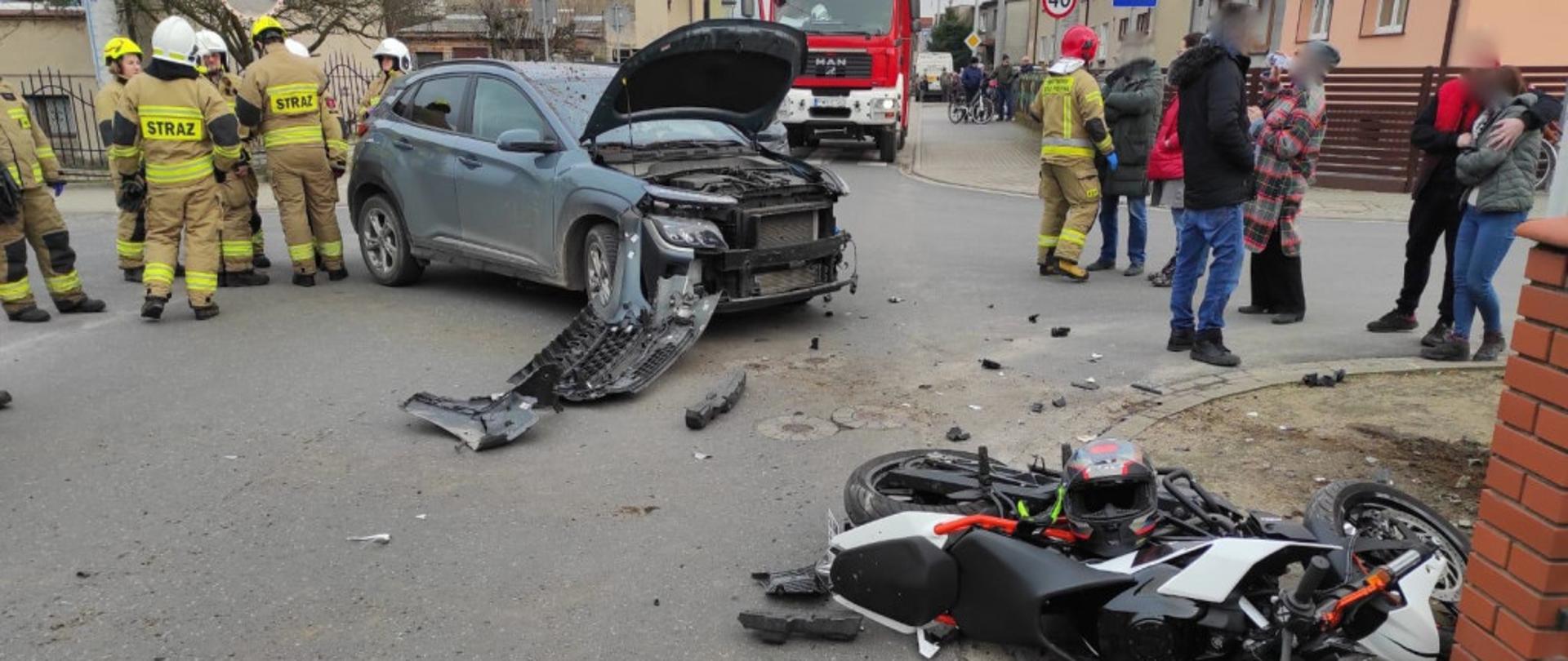 Zdjęcie przedstawia samochód osobowy i motocykl po zderzeniu na środku skrzyżowania. Przód samochodu uszkodzony, motocykl leży na chodniku. Po prawej stronie stoją świadkowie, po lewej stronie strażacy. Z tyłu wóz straży pożarnej. 