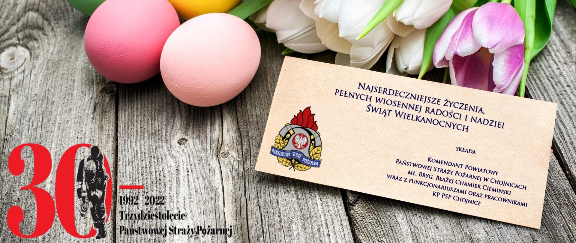 Na drewnianym blacie znajdują się tulipany, jajko oraz kartka z życzeniami Komendanta Powiatowego KP PSP w Chojnicach z okazji Świąt Wielkanocnych. 