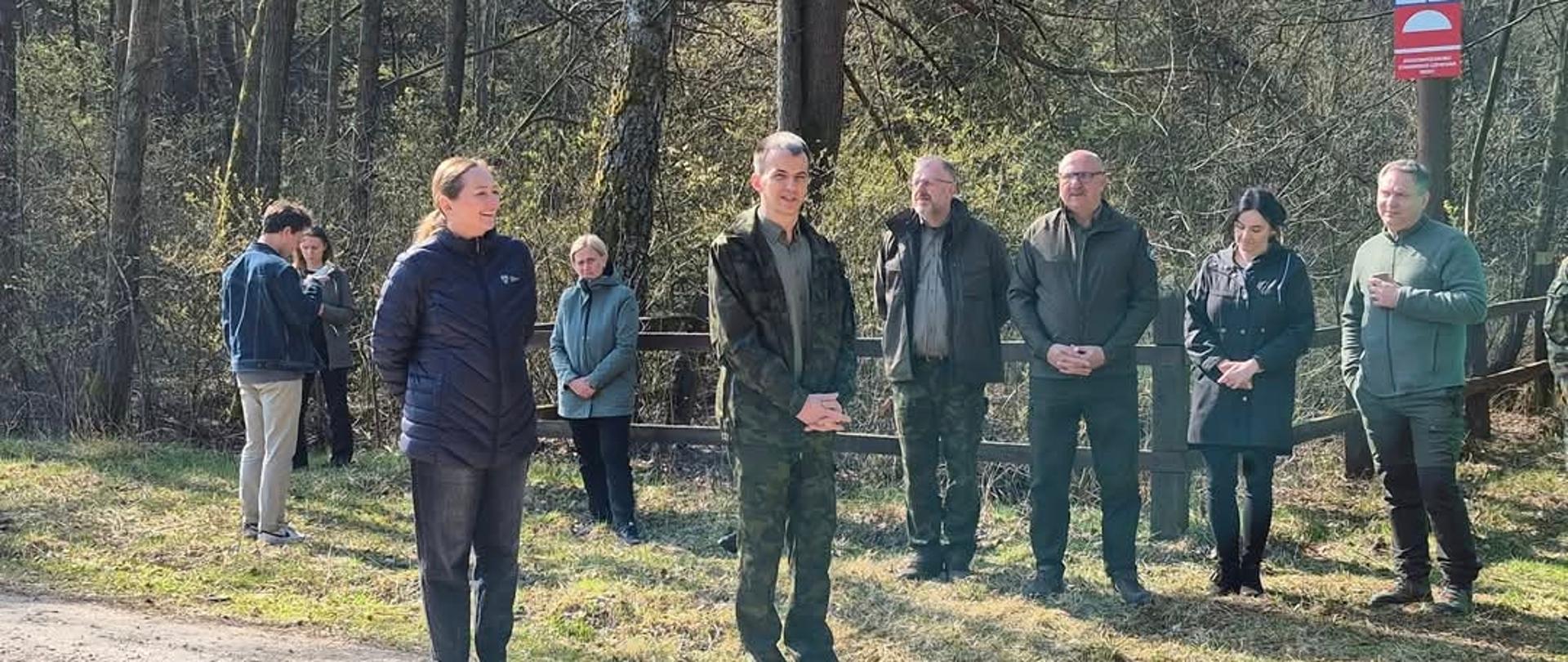 Zrzut ekranu z Facebooka przedstawiający zdjęcie grupy ośmiu osób stojących przy leśnej drodze w słoneczny dzień. Ludzie ubrani są w kurtki i odzież terenową, część stoi w szeregu, a w tle widać drzewa oraz tabliczkę przystanku. Pod zdjęciem widoczna nazwa profilu „Regionalna Dyrekcja Lasów Państwowych w Łodzi”.