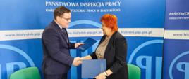 2 osoby na tle banneru w kolorze niebieskim z logo i napisem Państwowa Inspekcja Pracy. Osoby trzymają teczki w kolorze niebieskim i podają sobie dłonie w geście zawarcia umowy. Od lewej strony: Pan dr Karol Łapiński p.o. Okręgowego Inspektora Pracy w Białymstoku, Pani dr n. med. Maria Siewko – Podlaski Państwowy Wojewódzki Inspektor Sanitarny w Białymstoku.