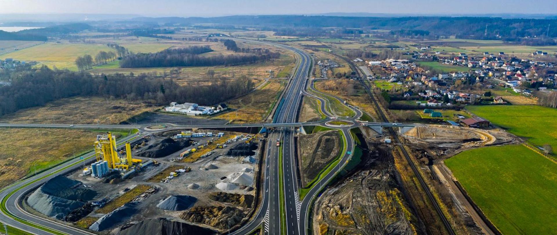 Widok z lotu ptaka na nowo wybudowany odcinek dwujezdniowej drogi ekspresowej z rondami i mostem. Po lewej stronie widać tereny przemysłowe i składowisko materiałów budowlanych, a po prawej pola uprawne oraz zabudowania miejscowości. Droga przecina otwarty, zielony krajobraz.