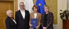 Na tle flagi Polski i UE stoją 4 osoby, 3 kobiety i 1 mężczyzna. Kobieta w beżowej marynarce trzyma granatową teczkę z logiem wojewody