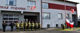 Na zdjęciu widzimy strażaków stojących w dwuszeregu oraz dowódcę i komendanta, po prawej stronie widzimy maszt na którym została zawieszona flaga Polski. W tle widzimy budynek komendy.