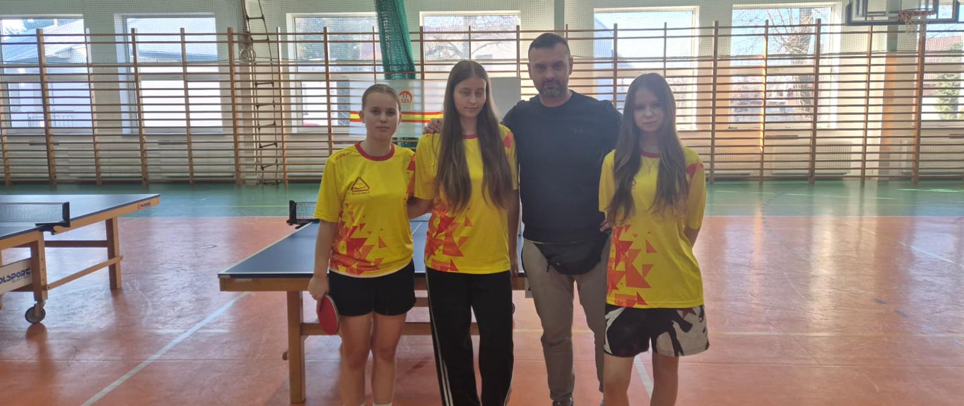 3 zawodniczki stojące z trenerem na tle stołu tenisowego
