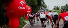 Na głównym planie kolarze biorący udział w wyścigu, z lewej strony czerwone balony z logiem sponsora