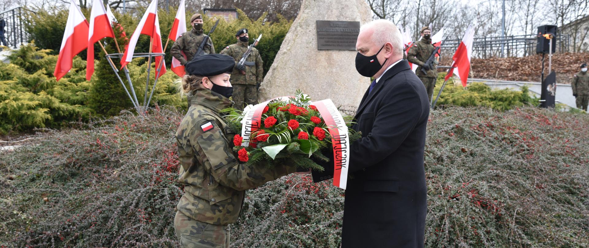 Wojewoda przekazuje żołnierce wieniec z biało-czerwonych kwiatów. W tle pomnik pamięci Żołnierzy Wyklętych (kamień z tablicą, na jego szczycie orzeł z rozpostartymi skrzydłami). Obok pomnika biało-czerwone flagi i warta honorowa żołnierzy 