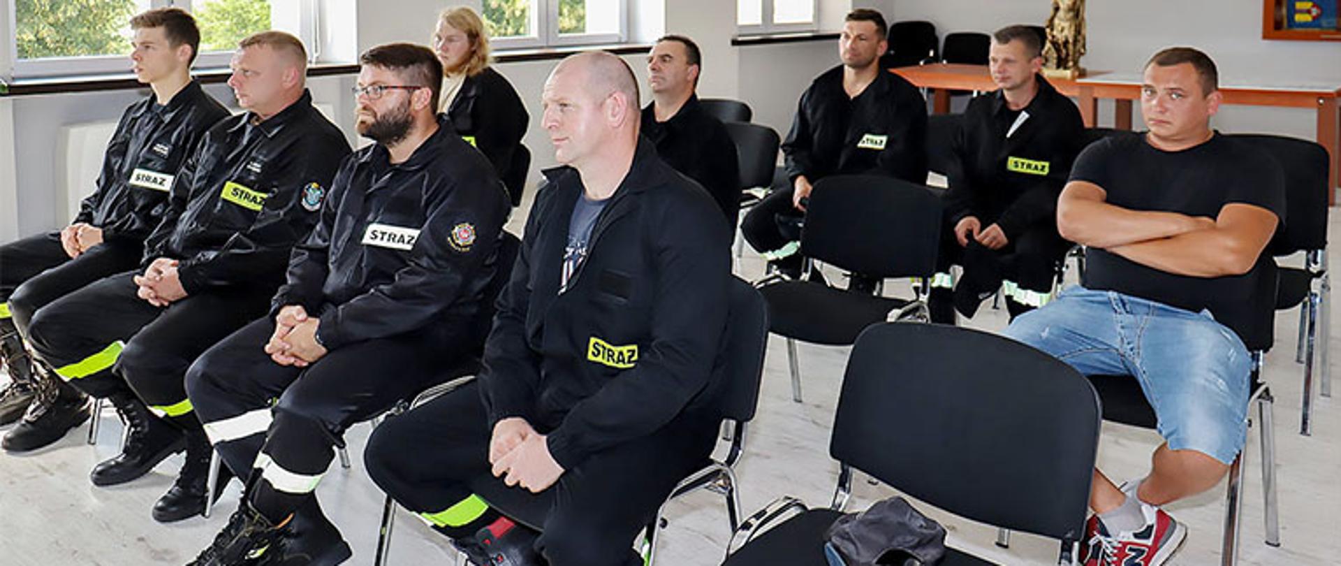 grupa dziewięciu druhów ochotniczych straży pożarnych ubrana w ubrania koszarowe barwy czarnej siedzi na krzesełkach w trzech rzędach, w tle widoczna figurka św. Floriana stojąca na stole, widoczny fragment sztandaru na ścianie osadzony w drewnianej gablocie, po lewej stronie trzy okna jedno u uchylonym skrzydłem, parkiet na podłodze w kolorze szarości