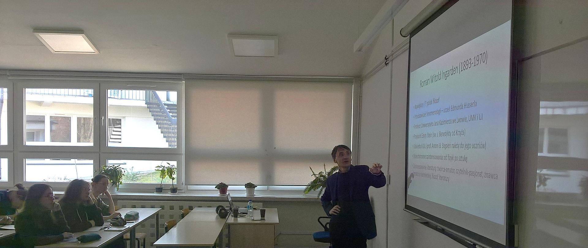 Jacek Wojtysiak prowadzi wykład z uczniami w sali lekcyjnej PLSP w Lublinie. Uczniowie siedzą w ławce. Na tablicy wyświetlany jest slajd przedstawiający ikonę.