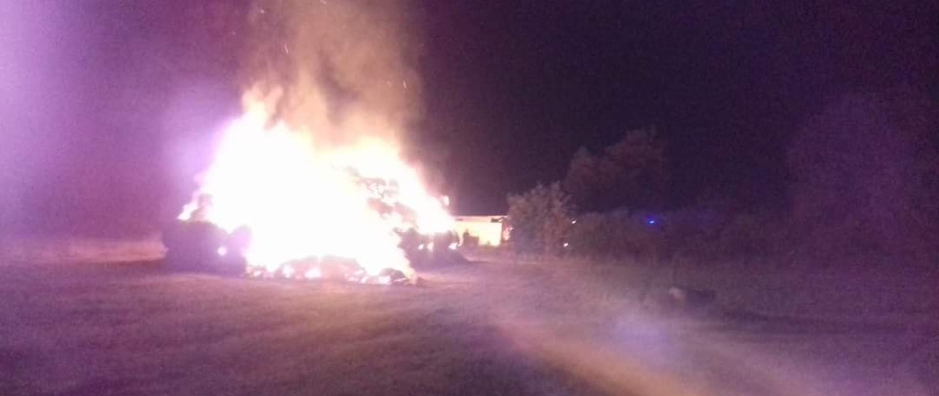 Na środku pola widać stóg słomy cały w ogniu, pora nocna, w oddali kilka drzew
