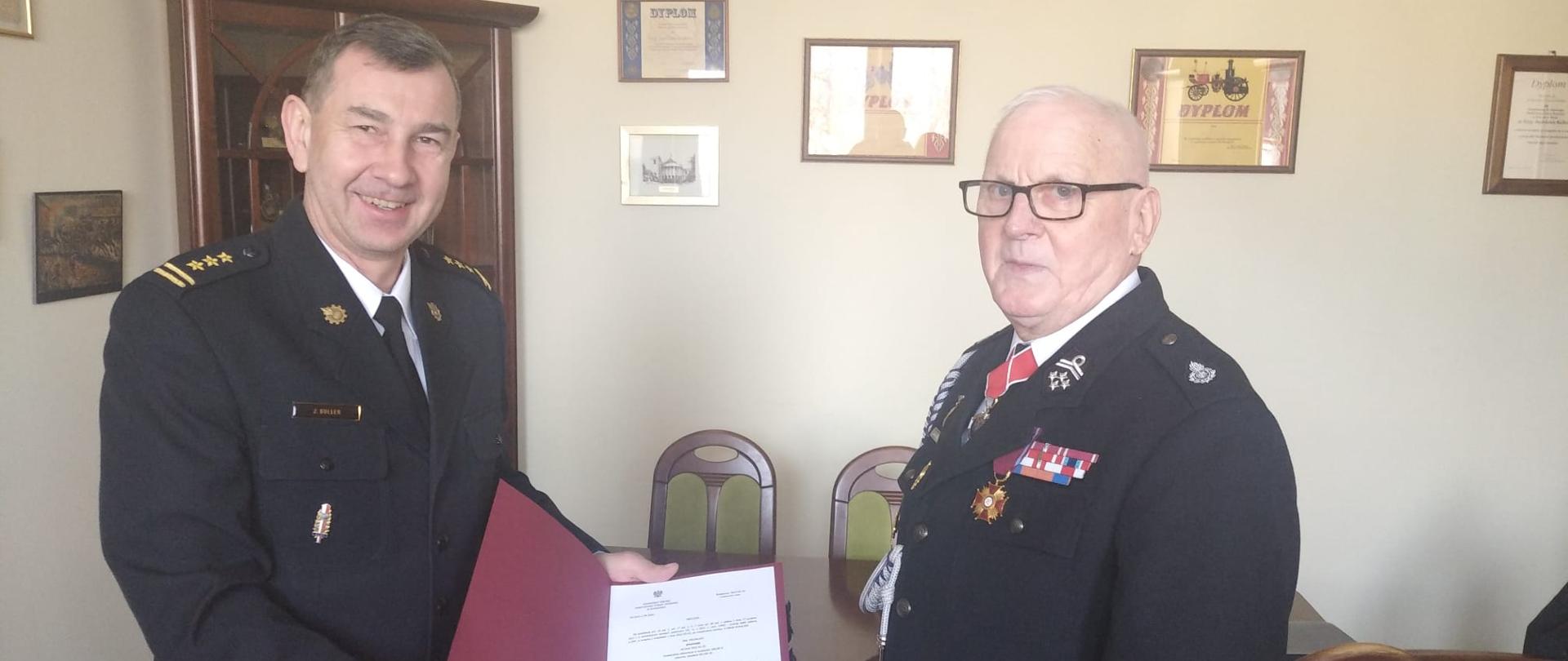 Z lewej strony Komendant Miejski PSP w Bydgoszczy st. bryg. Janisław Buller w granatowym mundurze wyjściowym PSP, z prawej druh OSP w granatowym mundurze wyjściowym OSP. Komendant trzyma otwartą teczkę koloru czerwonego z decyzją. Obydwaj znajdują się w biurze Komendanta, w tle kremowe ściany.
