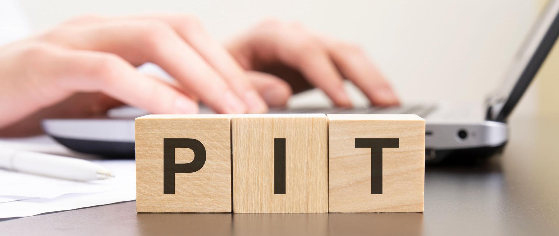 Klocki z literami PIT, w tle dłonie na klawiaturze komputera.