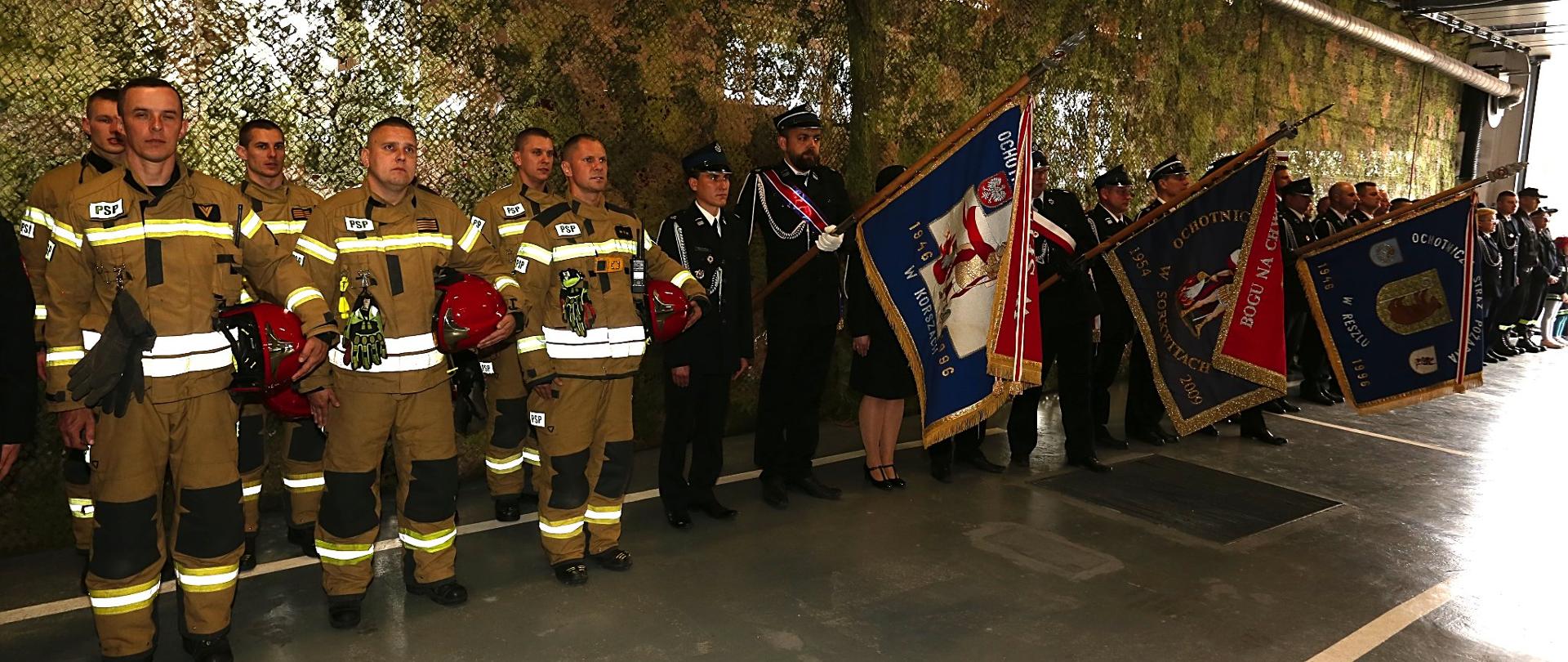 Zdjęcie przedstawia strażaków ustawionych w dwuszeregu na garażu JRG Kętrzyn. Od lewej strażacy ubrani w piaskowe Ubranie Specjale a pod lewą ręką trzymają hełm. Następnie druhowie z OSP z trzema sztandarami. W tle zielona siatka maskująca.