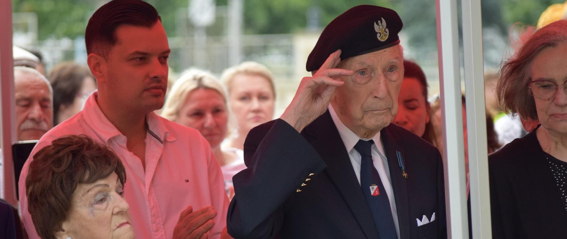 starszy mężczyzna w berecie salutuje przed flaga, wokół stoją inni młodsi ludzie 