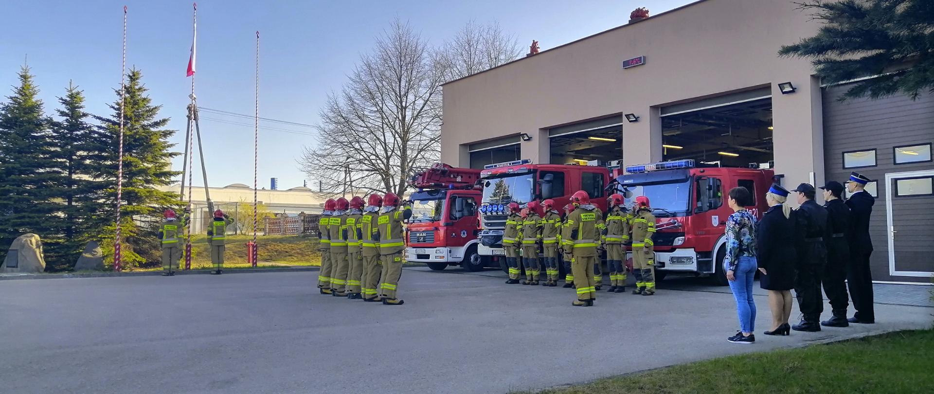 Zdjęcie przedstawia pododdziały strażaków państwowej straży pożarnej podczas podnoszenia na maszt flagi państwowej. Na drugim palnie zdjęcia widać samochody pożarnicze.