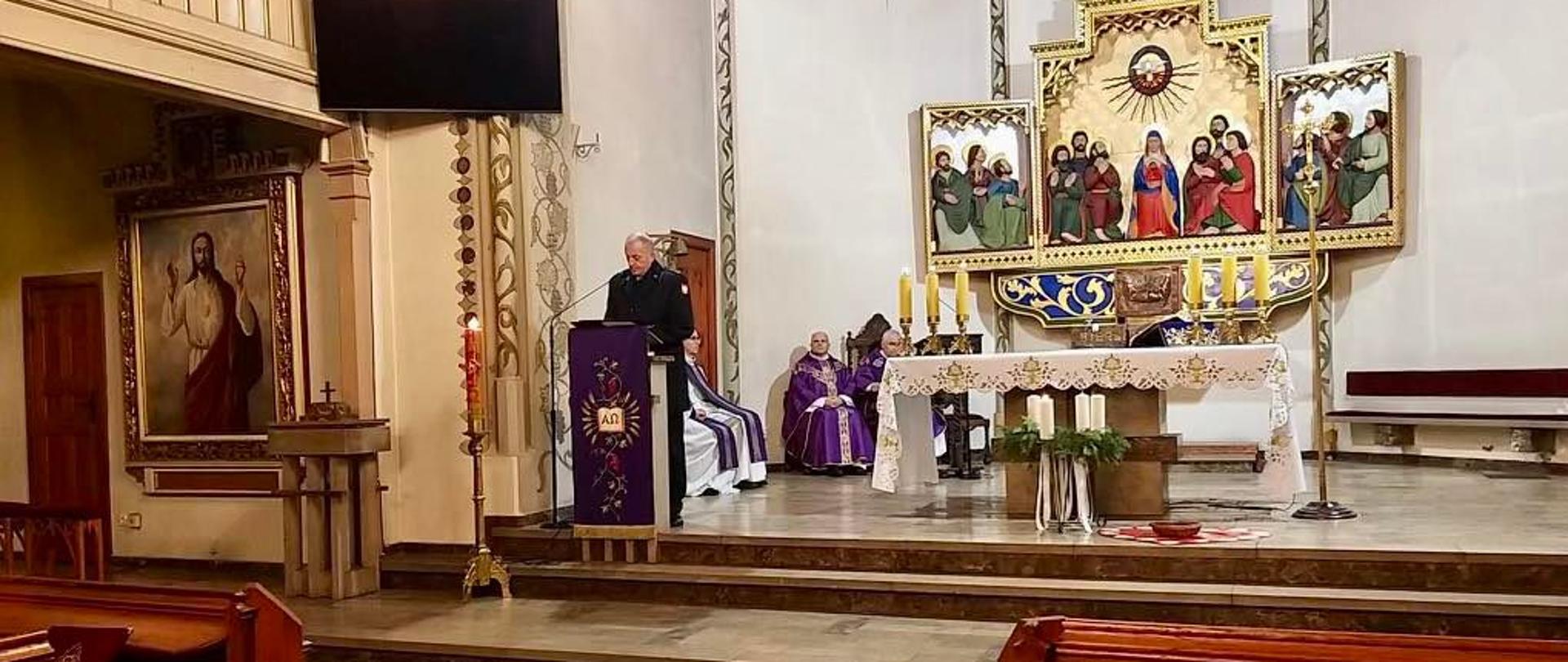Zdjęcie przedstawia ołtarz w kościele. Przed mównicą stoi i przemawia strażak w czarnym mundurze bez czapki. Za nim siedzi dwóch Księży. Na pierwszym planie widać ławki drewniana. W tle znajdują się piękne religijne obrazy. 