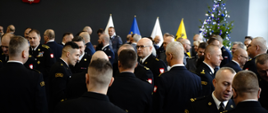 Strażacy oraz policjanci składają sobie życzenia na sali jest ubrana choinka na ścianie wisi godło Polski oraz stoją flagi.
