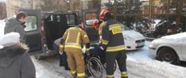 Zdjęcie przedstawia strażaków PSP pomagających starszej kobiecie na wózku inwalidzkim wjechać po najazdach do samochodu dostawczego zawożącego ją do punktu szczepień 
