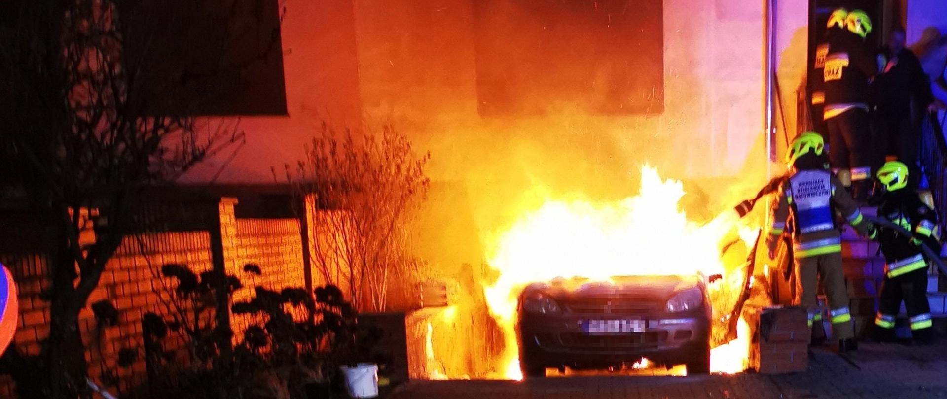 Widać palący się samochód na podjeździe przed budynkiem mieszkalnym, jest ciemno