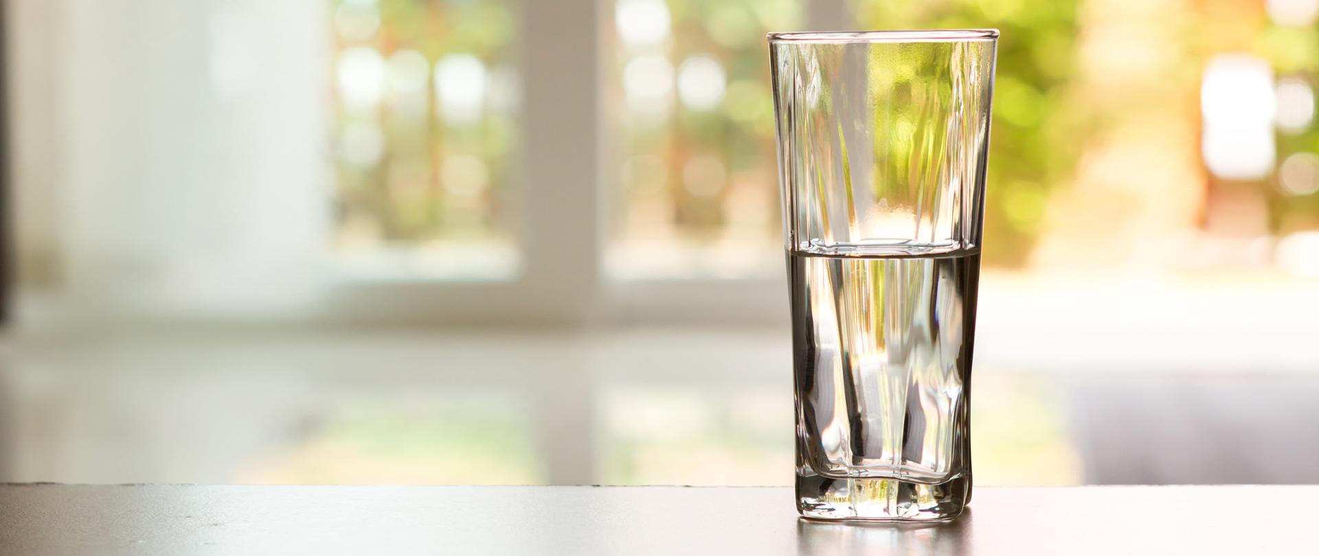 Wypełniona do połowy szklanka wody stojąca na blacie na tle rozmytego okna