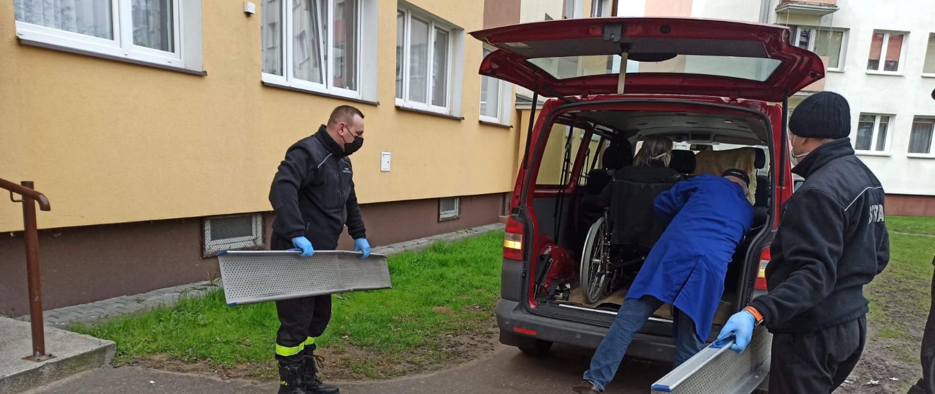 Zdjęcie przedstawia bordowy samochód typu bus stojący między pastelowymi blokami mieszkalnymi. W części bagażowej auta znajduje się człowiek na wózku inwalidzkim. Ubrany na niebiesko kierowca pojazdu odblokowuje urządzenie, a po bokach dwaj ubrani na czarno strażacy stoją w gotowości do podłożenia najazdów do wózka.