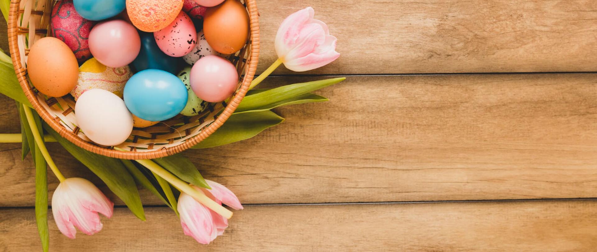 Grafika Wielkanocna - na drewnianym stole koszyk z kolorowymi pisankami, dookoła różowe tulipany