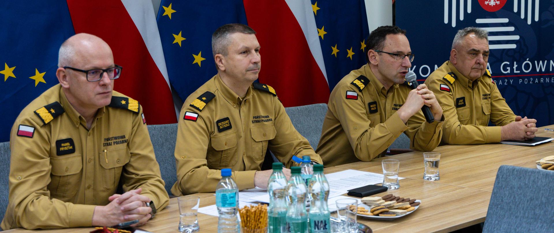Czterech funkcjonariuszy PSP w mundurach siedzi przy stole konferencyjnym, na stole znajdują się dokumenty, woda i przekąski, w tle flagi Polski i Unii Europejskiej oraz tablica z logo Komendy Głównej PSP.