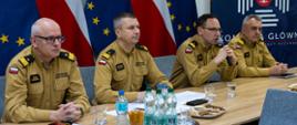 Czterech funkcjonariuszy PSP w mundurach siedzi przy stole konferencyjnym, na stole znajdują się dokumenty, woda i przekąski, w tle flagi Polski i Unii Europejskiej oraz tablica z logo Komendy Głównej PSP.