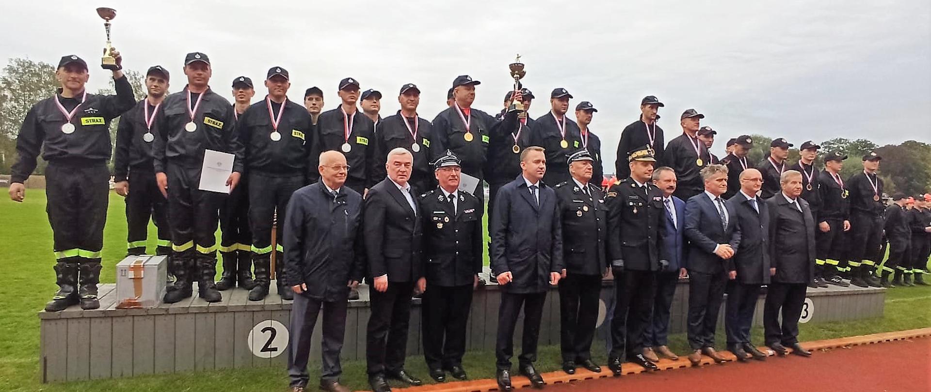 Zdjęcie przedstawia pamiątkowe ustawienie gości i organizatorów zawodów stojących na tle zwycięskich męskich drużyn ochotniczych straży pożarnych.
