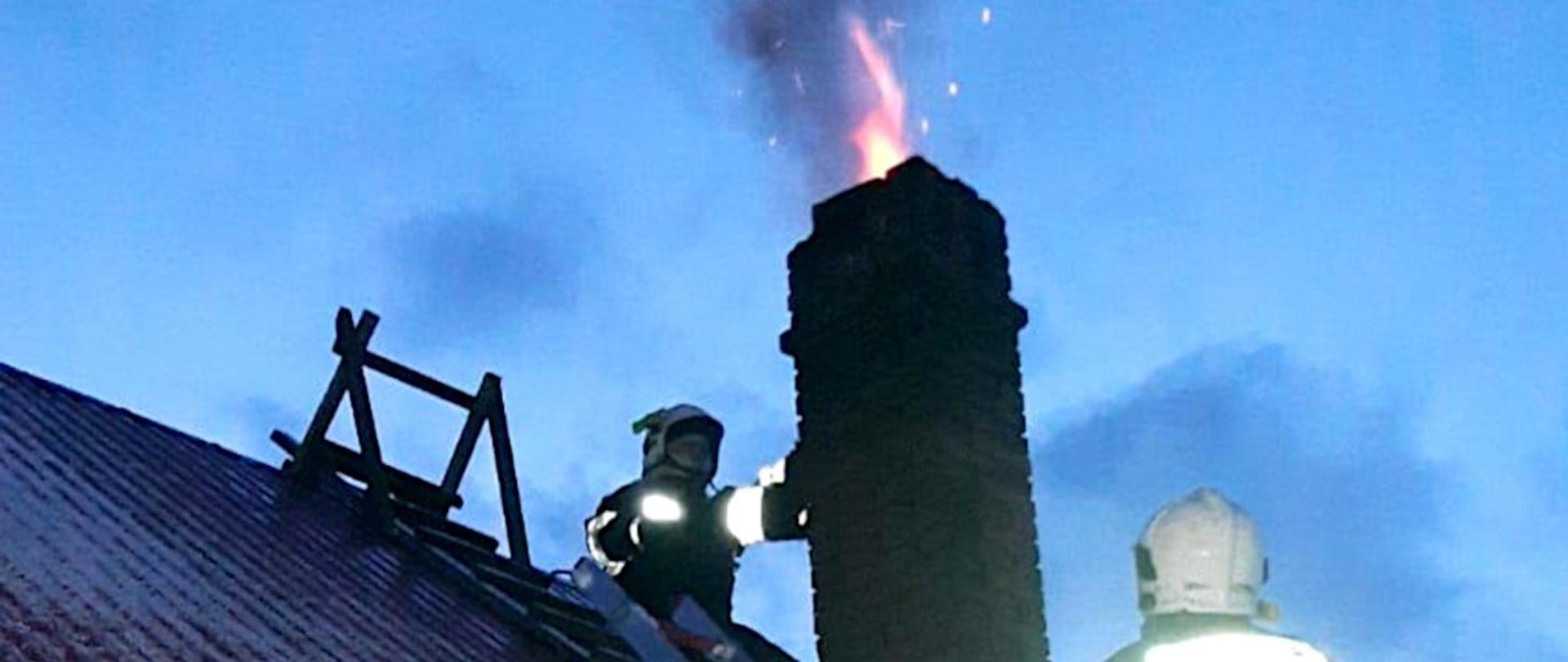 Na zdjęciu widoczny jest fragment dachu budynku i komin, z którego wydobywa się płomień z palących się sadzy. Obok komina dwóch strażaków w ubraniach specjalnych i hełmach.