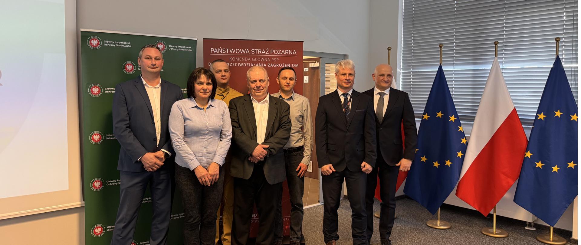 Grupa osób pozuje do pamiątkowego zdjęcia. Od lewej stoją:
Krzysztof Cząstkiewicz, Mazowiecki Wojewódzki Inspektor Ochrony Środowiska
Jolanta Odzimek, Główny Specjalista Wojewódzkiego Inspektoratu Ochrony Środowiska w Warszawie, Delegatura w Radomiu,
st. bryg. Łukasz Szymański, Komendant Miejski Państwowej Straży Pożarnej w Radomiu,
Tomasz Skuza, Kierownik Delegatury w Radomiu Wojewódzkiego Inspektoratu Ochrony Środowiska w Warszawie
Paweł Dadasiewicz, Naczelnik Wydziału Przeciwdziałania Poważnym Awariom Głównego Inspektoratu Ochrony Środowiska
Tomasz Osiński, Dyrektor zakładu SOUDAL Manufacturing sp. z o.o. w Pionkach
Michał Kacperski, Kierownik Działu R&D SOUDAL Manufacturing sp. z o.o.
W tle widać flagę Polski oraz Unii Europejskiej.
