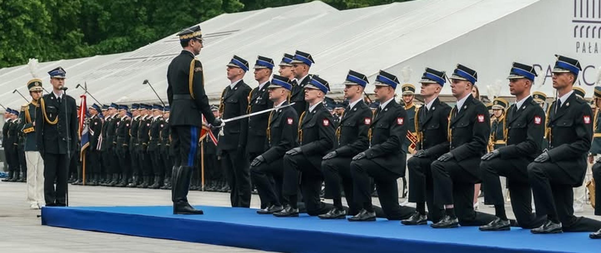 Strażacy podczas promocji oficerskiej klęczą na kolanie. Inny strażak szablą dokonuje promocji.