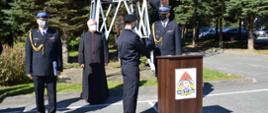 Komendant Powiatowy składa gratulacje strażakowi, który stoi w postawie zasadniczej. Obok stoi Ksiądz kapelan oraz Oficer w umundurowaniu galowym. Przed komendantem mównica.