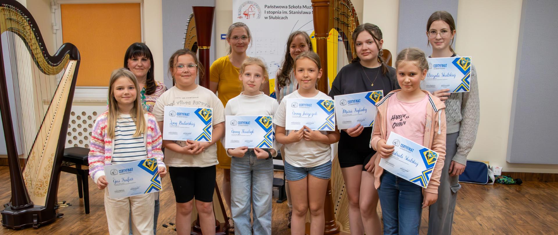 Osiem uczennic i trzy nauczycielki stoją w auli szkoły, dziewczyny trzymają w rękach dyplomy, za nimi stoją trzy harfy.