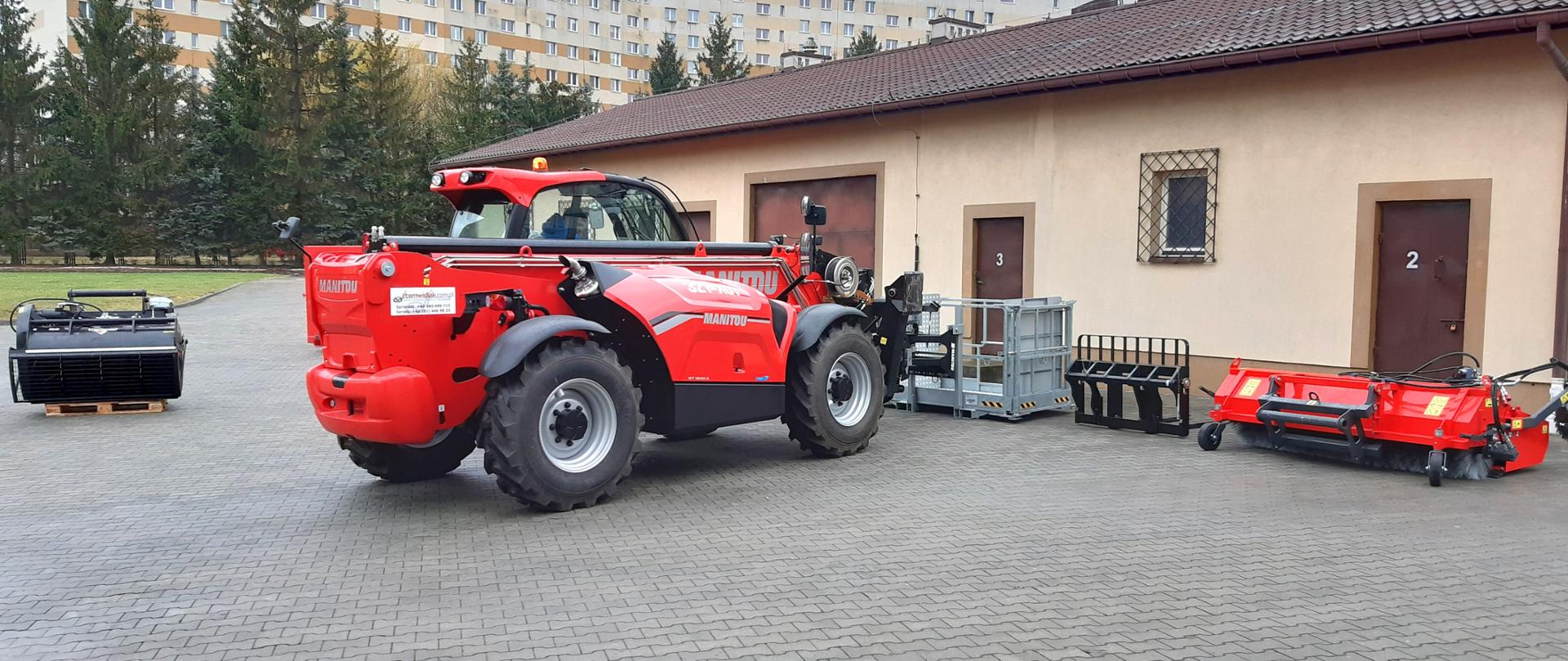 Zdjęcie przedstawia wielozadaniowy nośnik osprzętu marki Manitou znajdujący się na placu komendy miejskiej. W tle parterowy budynek magazynowy za którym znajdują się bloki mieszkalne. 