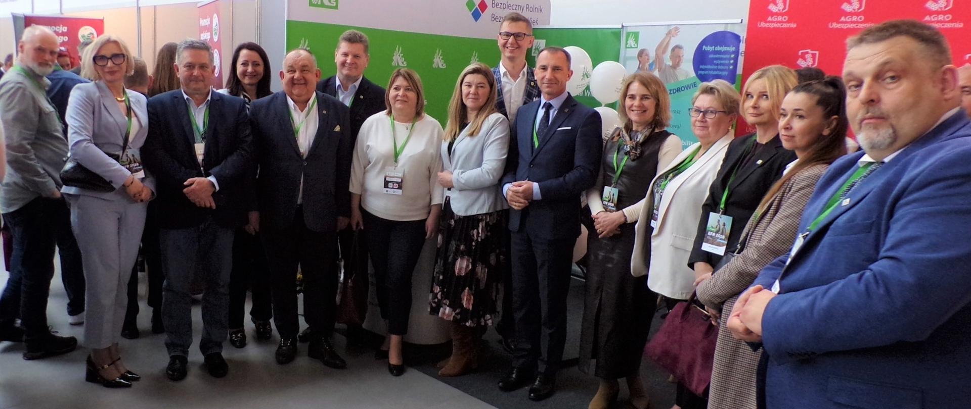 Kilkanaście osób pozuje do zdjęcia, stojąc przy stoisku informacyjno-promocyjnym Kasy.