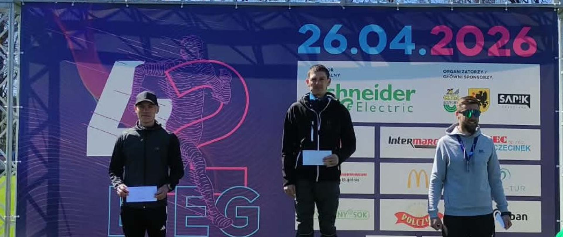 Trzech mężczyzn stoi na podium po biegu ulicznym w słoneczny dzień. Zwycięzca zajmuje środkowy najwyższy stopień z numerem 1, a pozostali stoją na miejscach 2 i 3 po bokach. W tle duży baner promujący wydarzenie z datą 26.04.2026 oraz logotypami sponsorów.